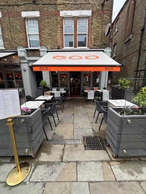 From sidewalk facing venue. Opening ahead between planters. Path behind between outdoors seating to entrance door on back.