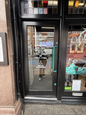 Glass door with black frames. Push green handle on right to open. One step up to door. Doormat behind. Assistance doorbell on left of door.