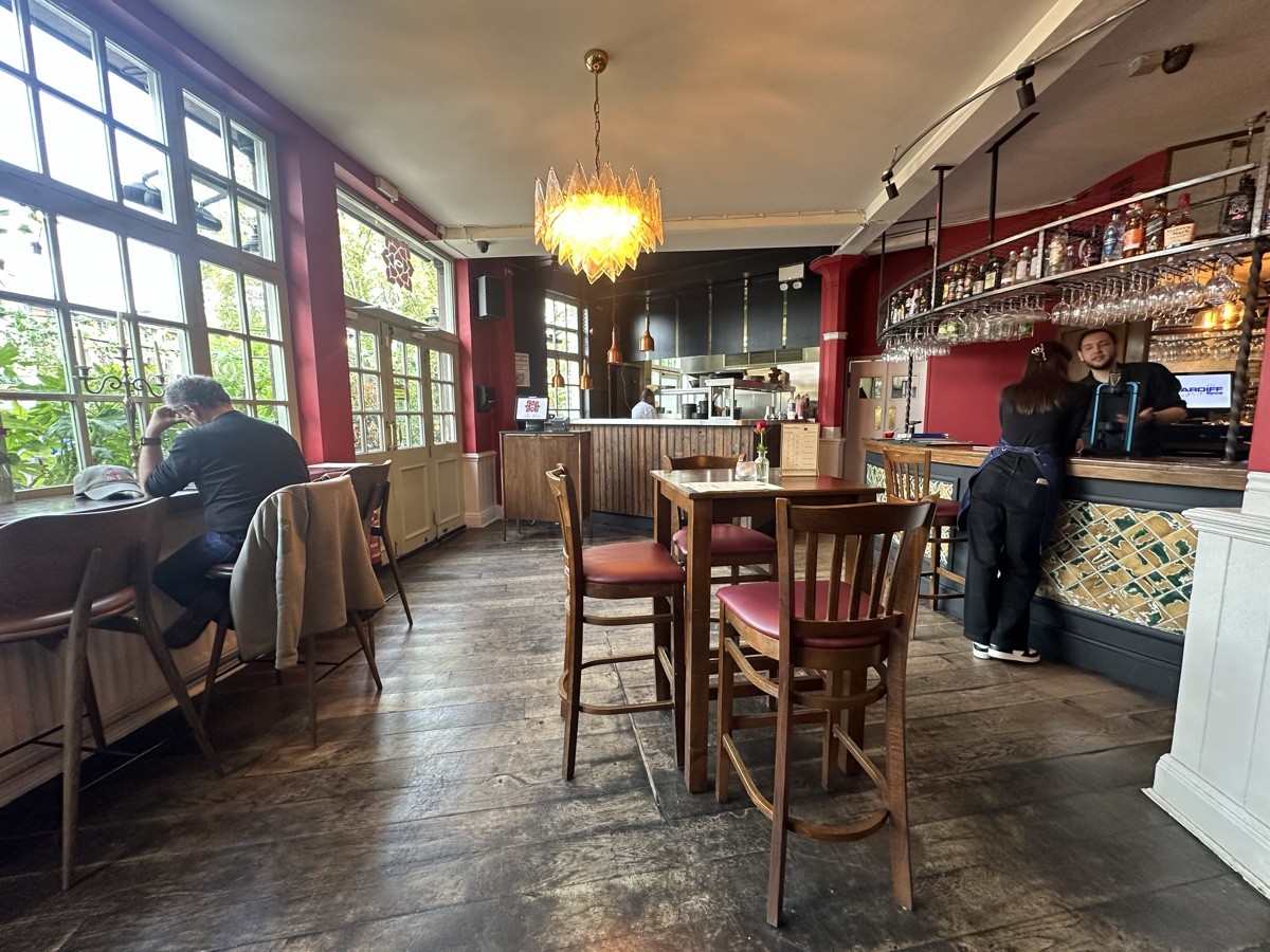 From main entrance facing left. High tables and stools to left and right. Open kitchen and alternative entrance behind. Bar to right.