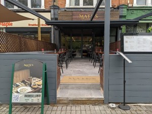 From pavement facing venue. A-frame sign on left, restaurant barrier behind on left and right. Two steps up ahead.