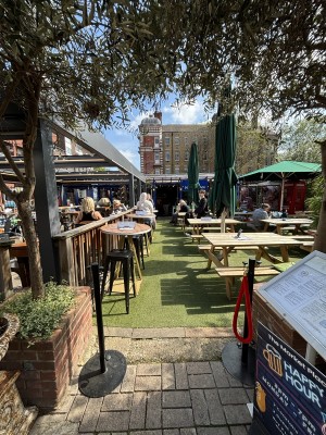 From pavement facing venue. Sign on right. Planters on left and right. Path in centre to entrance doors at the back. Outdoor seating ahead.