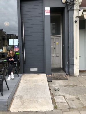 From pavement facing venue. Outdoor area to left. Ramp sloping upwards to entrance followed by door.