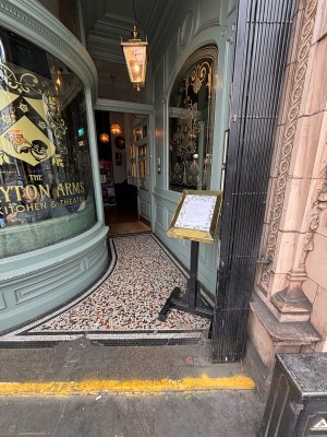 From pavement facing venue. One step up to black gate. Slide from left to right. Sign behind. Path ahead to door.