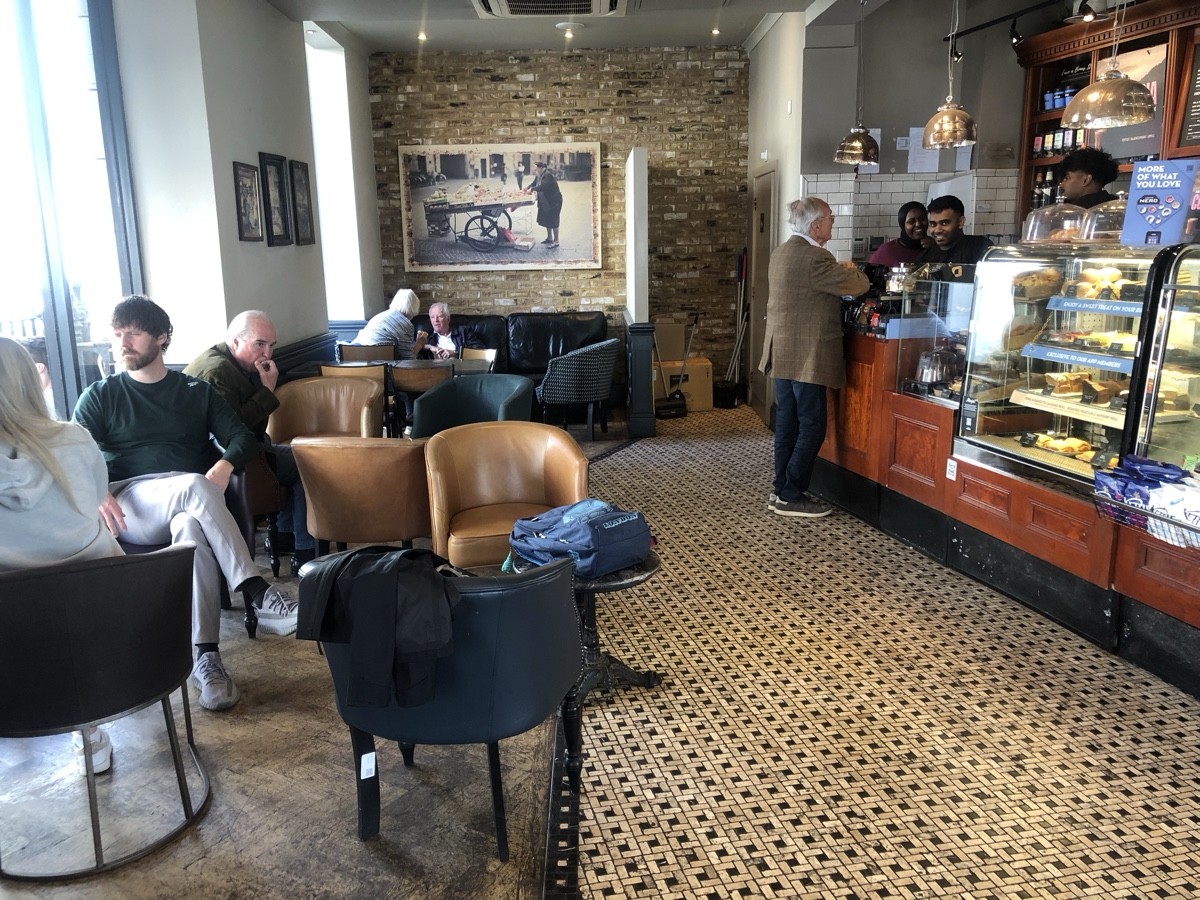 From entrance facing left. Tables with armchairs and chairs on left. Couch with table back left. Counter along right. Toilet on back right.