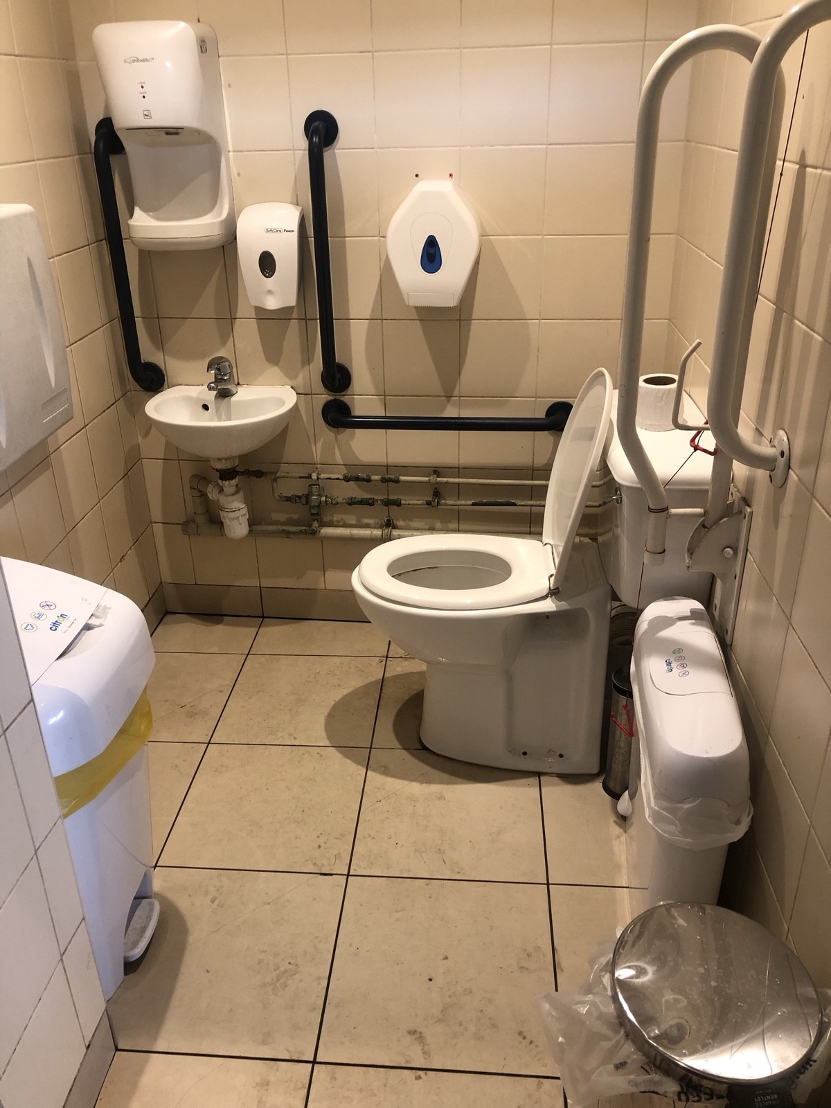 From bathroom door facing straight. Baby change table on back left. Toilet on right wall, emergency pull cord to right. Sink ahead on back left.