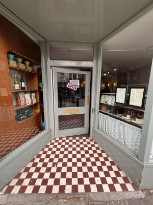 A sloped path ahead to door at the back. Glass door with grey frames. Push from right or press button on right to open.