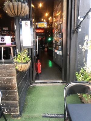 From pavement facing venue. Table with chairs on right. Slope upwards followed by step up and door.