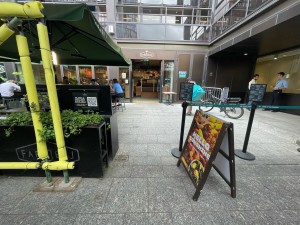 From sidewalk facing venue. Planters in left. Divider and sandwich board on right. Path straight in between leading to entrance in far back.