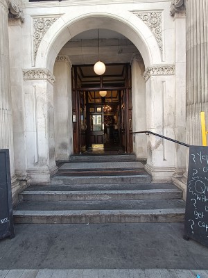 From street, facing venue. Three steps up to archway at back. A-frame board on right. Black, metal handrail along right.