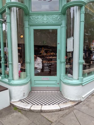 Glass door with teal frame ahead. Pull metal handle on right to open. Step up before door.