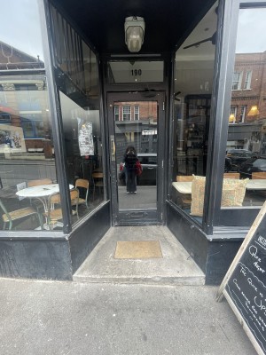 From street facing venue. Opening with step up ahead. Glass door with black frame at back. Pull gold metal handle from left to open.