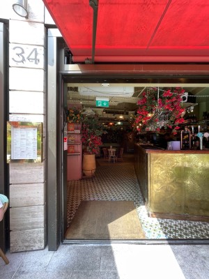 Facing entrance. Metal framed doorway ahead. Doors left open during opening hours. Seating on either side of entrance.