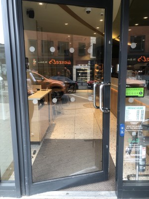 Glass door with black frame. Push silver metal handle on right to open. Doorbell for assistance to right of door.