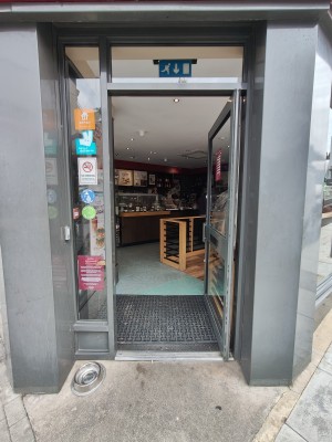 Grey framed glass door. Push grey handle on left to open. Doorbell for assistance on left.
