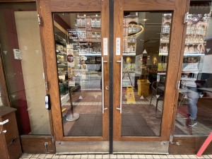 Wood framed glass doors ahead. Pull from metal handles in centre to open. Assistance bell on left.