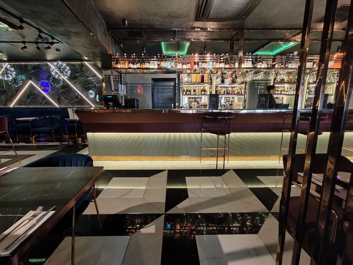 From column facing straight. Table and chairs on left. Path in centre to bar and stools at back. Second metal partition on right.