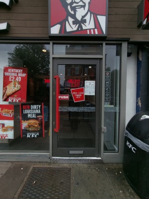 Black framed glass door. Push red handle on left to open. Doorbell for assistance on right of door.
