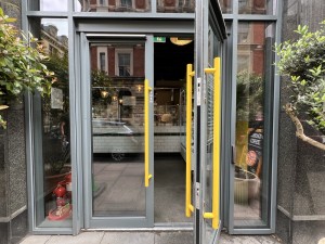 Grey and glass panelled door ahead. Push from metal handles in centre to open. Push button to right of door to open.