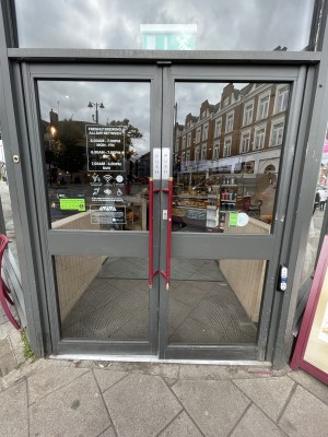 Grey framed glass doors. Push red handles in centre to open. Doorbell for assistance on right of doors.