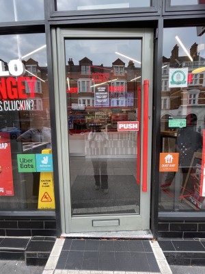 Facing glass door with gray frame. Push bright red handle on right to open.