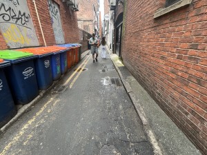 From corner of side street facing straight. Rubbish bins to the left. Sidewalk to right against wall. Door to staff area to centre back on right.