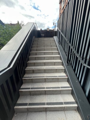From bottom of stairs facing straight. Staircase leads up to outdoor landing. Grey handrails on boths sides.