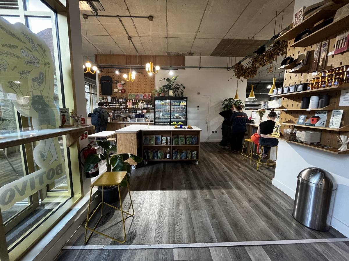 From door facing left. High table & stool on left. Product counter and fridge back. Toilet to its right. Product shelf on right. High tables behind.