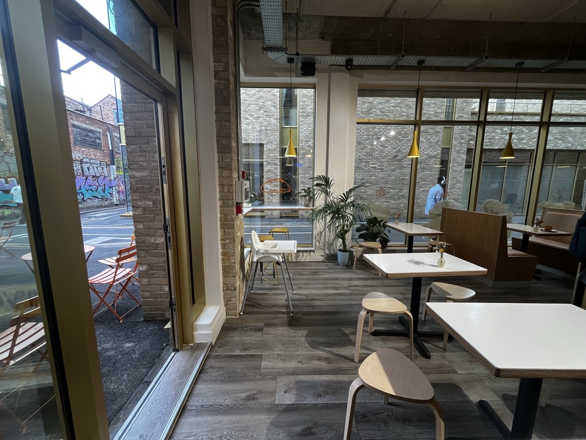 From product counter facing left. Pillar back left. High table and stools behind. Tables and stools on right. Booths behind.