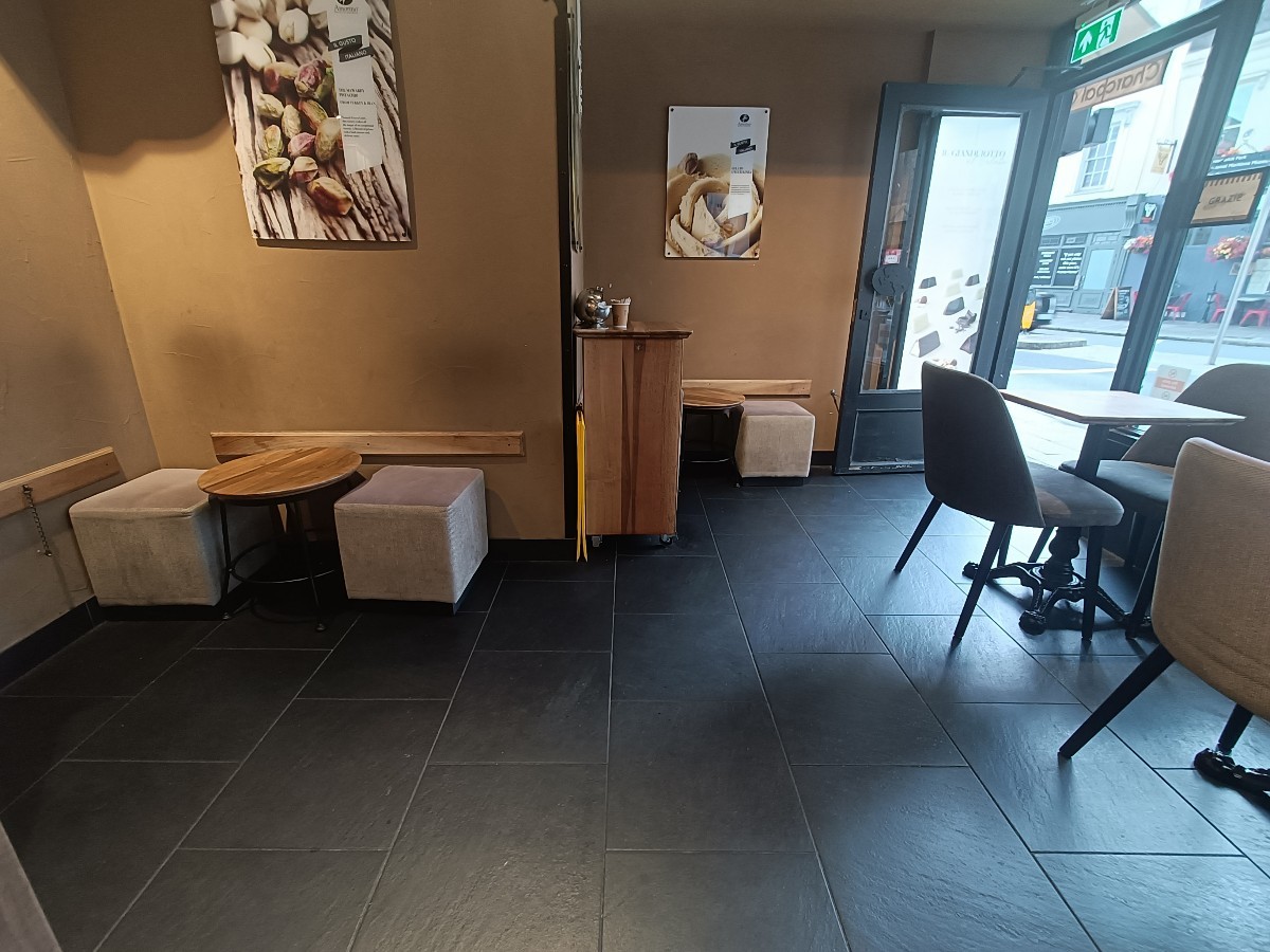 From opening, facing straight. Table and stools at back left. Condiments station at back. Tables and stools behind. Tables and chairs along right.