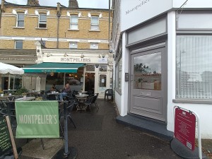 From street, facing venue. Restaurant barriers on left. Outdoor seating behind. Path ahead to doorway at back.