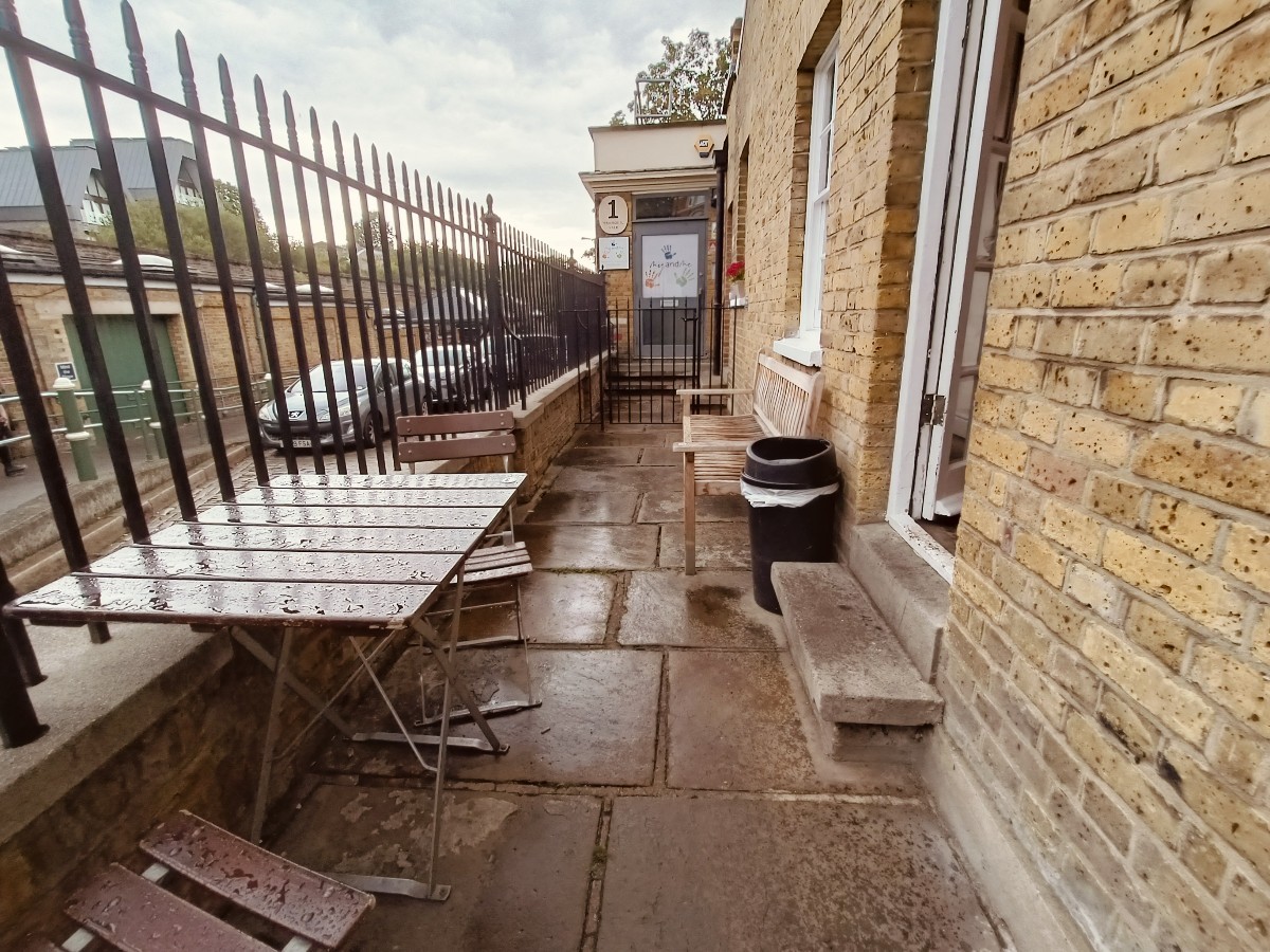 From end of tables and chairs, facing straight. Tables and chairs in left. Steps and doorway on right. Bin behind. Bench beyond.