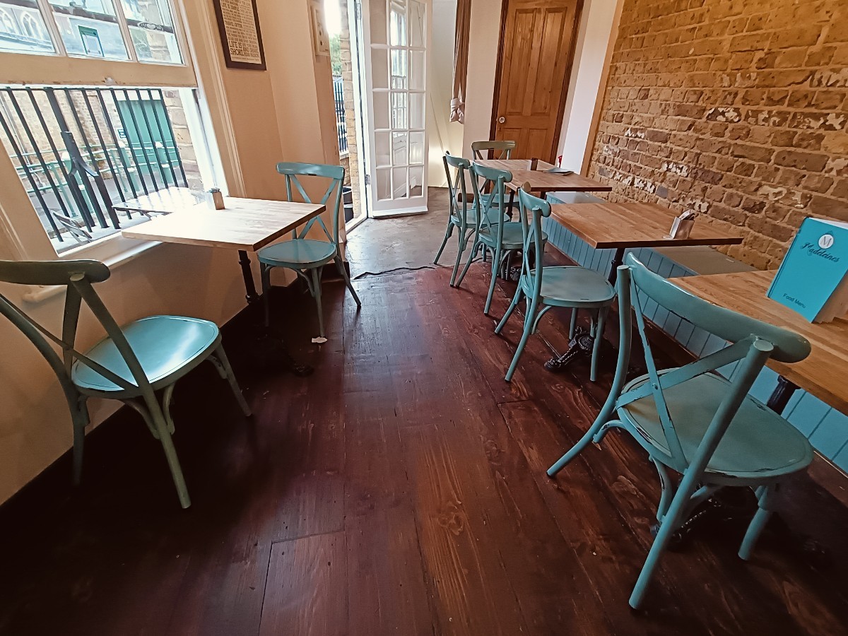 From end of counter, facing straight. Tables and chairs on left and along right. Doorway to outdoor at back left. Toilet at back right.