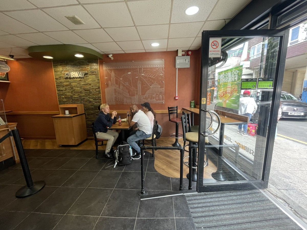 From entrance facing right. Metal barriers ahead. High table and stools along right. Tables and chairs at back.