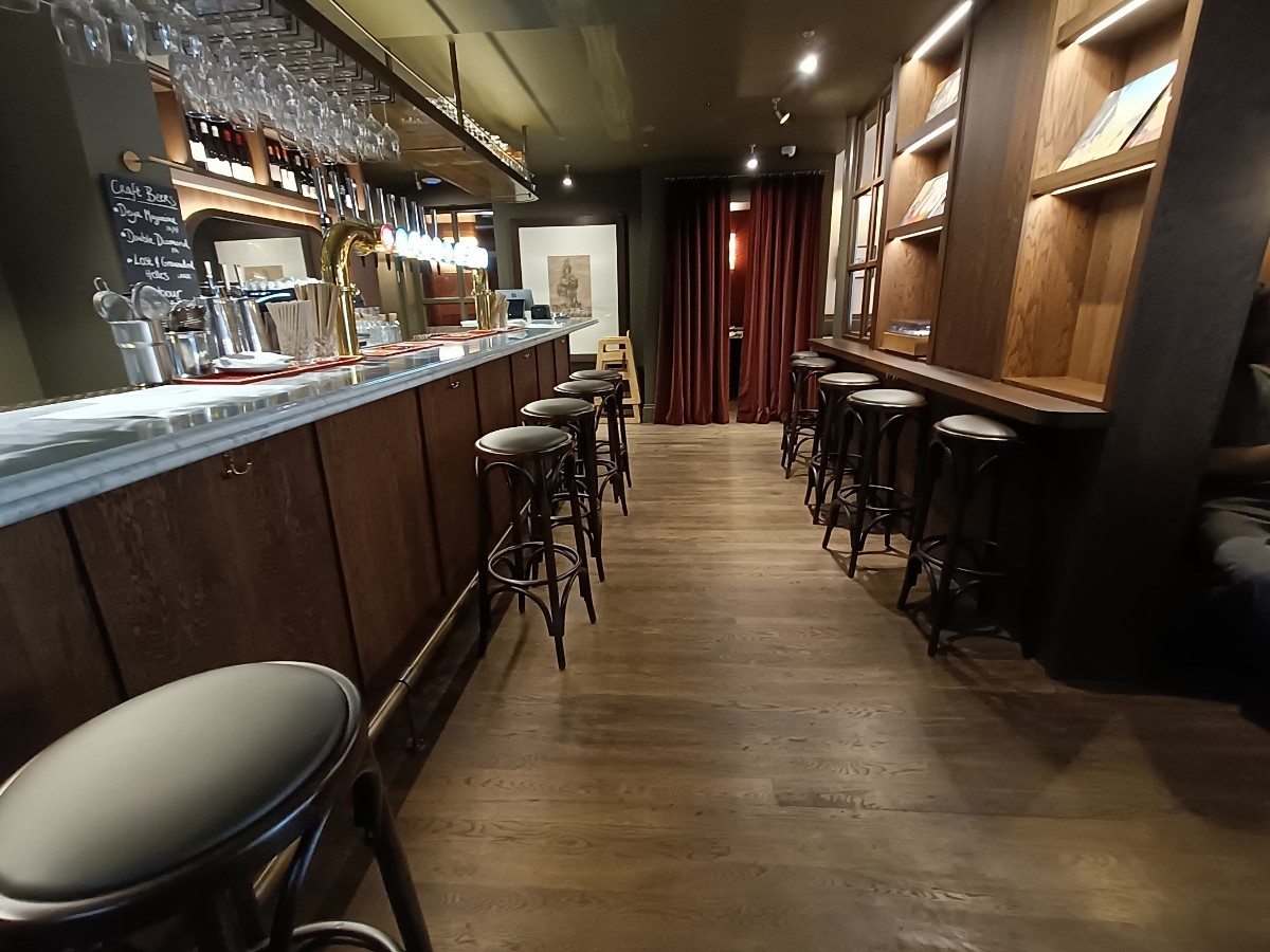 From bar counter, facing right. Bar counter and stools along left. High tables and stools along right.