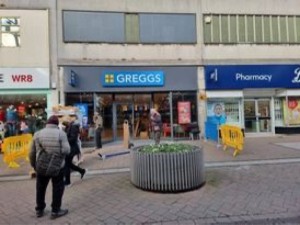 Photo shows Greggs taken from a distance. The shop is in the background and the high road and a planter is in the foreground.