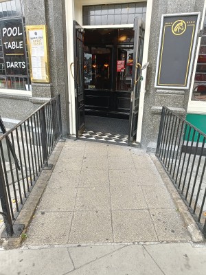 From street, facing venue. Ramp up ahead to doorway. Black, metal handrails along both sides.