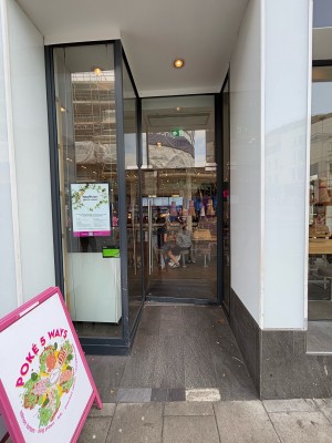 From sidewalk facing venue. Opening ahead. Glass door with black frame on back. Push by a metal handle on left to open.