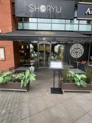 From sidewalk facing venue. Opening ahead between planters. Standing sign on right. Path behind with seating along both sides to entrance on back.