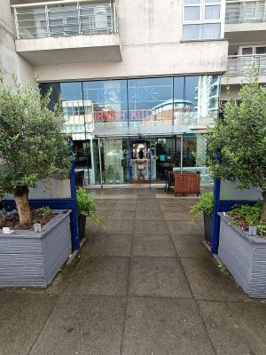 From street, facing venue. Opening ahead. Planters on both sides. Outdoor seating behind.