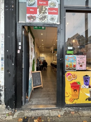 From sidewalk facing venue. Black wooden door with advertising on the front. Glass wall to the right with colorful posters
