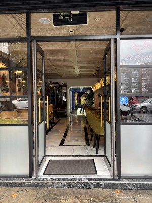 From sidewalk facing straight. Glass doors with gold handles on each side. Doormat at foot of doorframe.