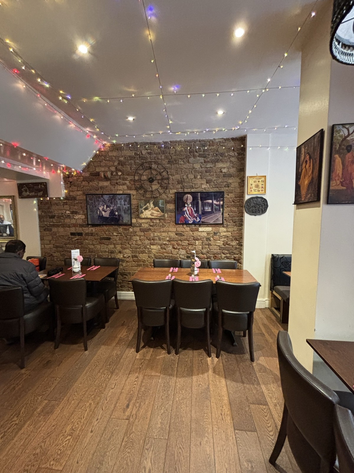 From midsection facing right. Tables and chairs, brick wall at the back with picture frames. Column to the right