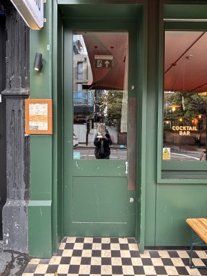 From sidewalk facing entrance. Green wooden door with glass centre. Menu on left hand side attatched to column