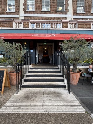 From sidewalk facing straight. Steps leading to main entrance. Black handrails on each side. Black and white steps. Plants on each side of stairs.