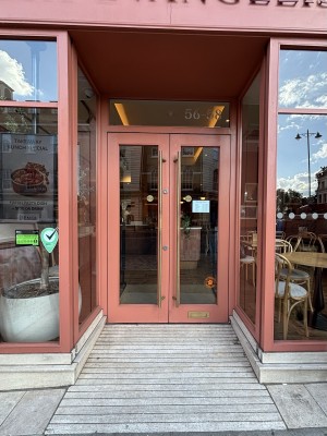 From sidewalk facing straight. Pink doorframe with glass centre. Golden door handles on each side. Pull to open. Ramp leading to entrance
