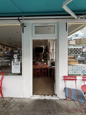 From sidewalk facing straight, white doorframe with glass centre, metal door handle to the left, push to open. Large glass windows on each side