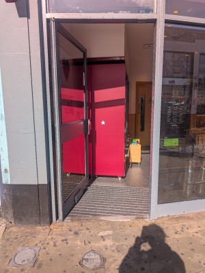 Grey frame door. Metal handle on right. Push to open. Bump at entrance. Doormat after.