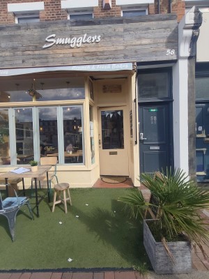 From street facing venue. Planter decor on right. Grass path down centre to entrance on back. Outdoor seating on left.