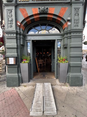 From sidewalk facing venue. Opening ahead with planters on the sides. Green wooden doors with glass panels on back. Push from center to open.
