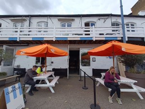 From street, facing venue. Picnic tables with parasols on both sides. Doorway at back. Queue cord on right.